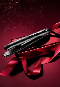 Schwarzer Haarglätter mit glatten Platten und metallischen Akzenten, auf tiefrotem Geschenkpapier und Schleifen liegend, mit glänzender Oberfläche.