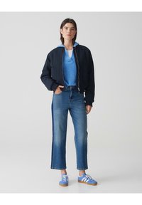 Marineblaues Bomberjackett über einem hellblauen Pullover, kombiniert mit geraden Denim-Jeans und blauen Sneakers mit weißen Streifen.