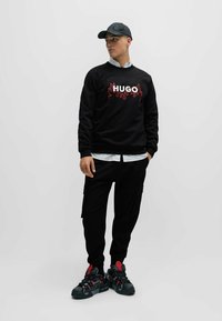 Sudadera negra con "HUGO" en llamas blancas y rojas, combinada con pantalones cargo negros y zapatillas robustas con acentos rojos y suelas negras.