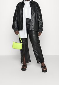 Svart läderjacka i oversize-stil över vit skjorta, kombinerad med svarta läderbyxor med sidofack, kraftiga svarta sandaler och en neongrön handväska.