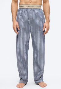 Ανδρικό παντελόνι πιτζάμας με κάθετες γκρι και λευκές ρίγες, με μπεζ λάστιχο μέσης με το λογότυπο Calvin Klein, φορεμένο ξυπόλητο σε λευκό φόντο.