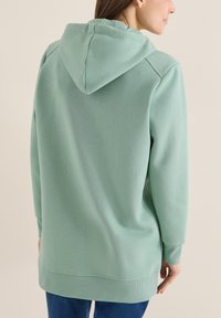 Femme portant un sweat à capuche surdimensionné vert clair, vue de dos, sur un fond beige uni.