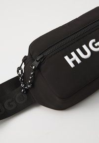 Sac en tissu noir avec fermeture éclair, arborant un logo blanc "HUGO" bien en vue et une bandoulière texturée avec des accents noir et blanc.