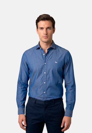 Uomo con capelli castani corti, indossa una camicia blu abbottonata e pantaloni scuri, in piedi di fronte a uno sfondo semplice.