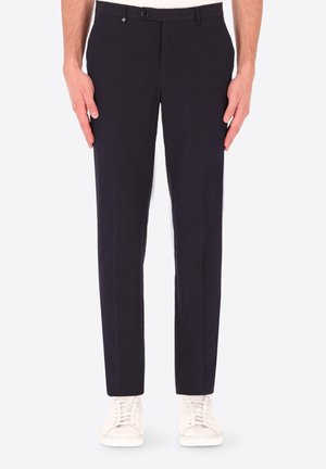 Mann trägt schwarze maßgeschneiderte Hose und weiße Turnschuhe, steht gerade vor einem einfarbigen weißen Hintergrund.