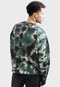 Mand iført en grøn og sort tie-dye sweatshirt, står sidelæns med ryggen synlig, parret med sorte jeans foran en ensfarvet baggrund.