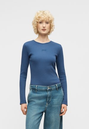 Jeune femme aux cheveux blonds bouclés et courts, portant un haut bleu à manches longues de la marque GAP et un jean bleu taille haute, devant un fond clair uni.