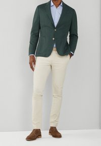 Tmavě zelený blazer s špičatými klopami, dvěma knoflíky a předními kapsami, spárovaný se světle modrou košilí, béžovými slim-fit kalhotami a hnědými botami.
