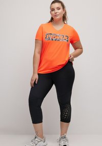 Oranje sportt-shirt met een zwart-wit 'POWER' grafiek, gecombineerd met zwarte 3/4 leggings met mesh uitsparingen en textuurdetails.