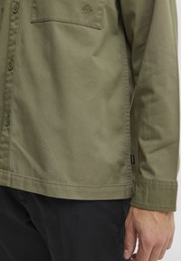 Olive green button-up shirt with a chest pocket, flat collar, and visible stitching details, paired with black pants.