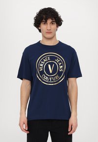 Tumši zila kokvilnas t-kreklu, ar lielu, zeltītu apļa logotipu, uz kura redzams teksts "VERSACE JEANS COUTURE" un centrālais "V" dizains.