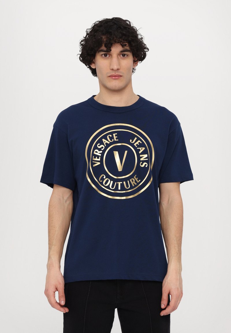Tumši zila kokvilnas t-kreklu, ar lielu, zeltītu apļa logotipu, uz kura redzams teksts "VERSACE JEANS COUTURE" un centrālais "V" dizains.