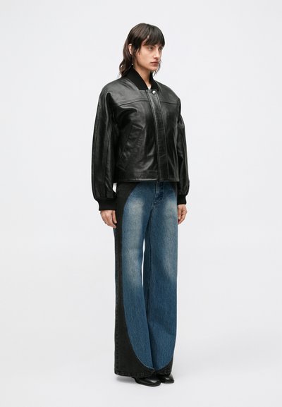 Μαύρο δερμάτινο μπουφάν bomber με ριμπ γιακά, συνδυασμένο με τζιν φαρδιάς γραμμής με σχέδιο χρωματικών μπλοκ σε αποχρώσεις του μπλε και μαύρου.