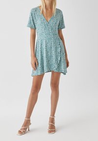 Femme portant une robe à fleurs teal à manches courtes et décolleté en V avec des sandales à talons hauts blanches, se tenant devant un fond uni.