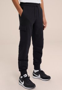 Svarta cargobyxor med elastisk midja, sidofickor, texturerat tyg och ribbad benslut, tillsammans med svarta Nike-sneakers.