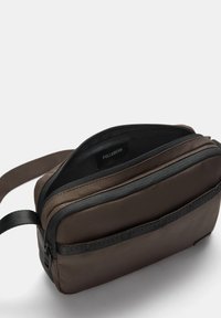 Braune Stofftasche mit strukturierter Oberfläche, die zwei mit Reißverschluss versehene Fächer, schwarze Reißverschlussakzente und einen verstellbaren Tragegurt aufweist.