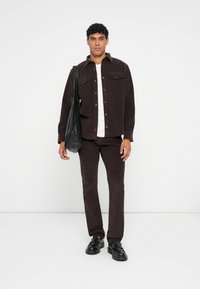 Camisa de pana marrón y pantalones a juego, camiseta blanca debajo, bolso negro cruzado, zapatos negros, de pie frente a un fondo liso.