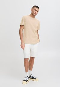 Beige strukturierte Kurzarmbluse kombiniert mit weißen Shorts. Schwarze und beige Sneakers mit weißen Schnürsenkeln, die vor einem neutralen Hintergrund stehen.