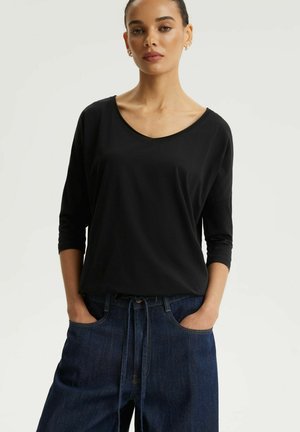 Kobieta ubrana w czarną bluzkę z dekoltem w kształcie litery V i rękawami o długości do połowy przedramienia oraz luźne, ciemnoniebieskie spodnie dżinsowe z rękami w kieszeniach na jednolitym tle.