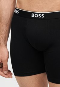 Αρσενικός κορμός που φοράει μαύρα boxer briefs με το λογότυπο "BOSS" στη μέση, το χέρι χαλαρό δίπλα στο σώμα, δέρμα ορατό πάνω και κάτω από το ένδυμα.