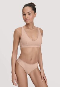 Femme portant un soutien-gorge en dentelle beige et un shorty assorti, se tenant devant un fond gris clair, regardant droit devant.