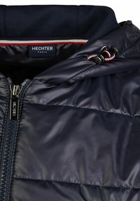 Donkerblauwe gewatteerde jas met ritssluiting, capuchon met trekkoord en voering met rood-witte accenten aan de binnenkant. Merketiket zichtbaar aan de binnenzijde.