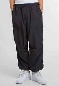 Zwarte, lichtgewicht, loszittende broek met een elastische tailleband en trekkoord. Zijzakken en taps toelopende boorden. Draagt ze met witte sneakers.