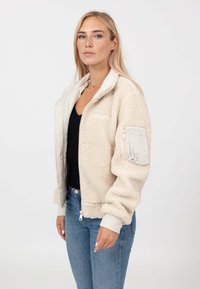 Giacca in pile beige con zip frontale, polsini a costine e fodera interna trapuntata. Presenta una tasca a toppa sul maniche sinistro.