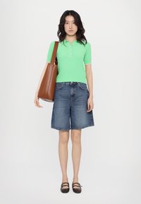 Green knit polo shirt with short sleeves and button detail, paired with denim Bermuda shorts. Brown tote bag and black strap shoes visible.
