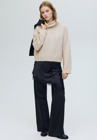 Bézs színű kötött pulóver magas gallérral, hozzá fekete szatén csipkés szegéllyel díszített szlip ruha és széles szárú fekete nadrág. Fekete bokacsizma.