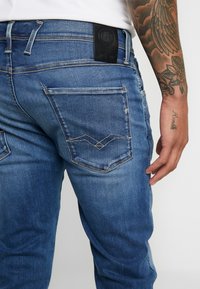 Jeans din denim albastru au o textură ușor decolorată, cusături în contrast și un etichetă din piele neagră pe talie.