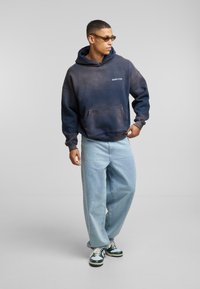 Felpa con gradient navy e grigio, dotata di tasca frontale, abbinata a jeans loose-fit blu chiaro e sneakers con dettagli verdi.