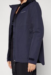 Veste bleu marine imperméable avec fermeture éclair, capuche ajustable et sangles de poignet en Velcro. Présente une texture mate et un design simple.
