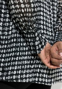 Sort bluse med hvidt og gråt abstrakt polkaprikket mønster. Gennemsigtige ærmer og knapmanchetter, fremstillet i blødt, struktureret stof.