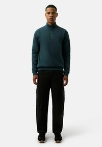 Ciemnozielony polar z wysokim kołnierzem i ćwierć zamkiem, zestawiony z luźnymi czarnymi spodniami cargo i czarnymi trampkami.