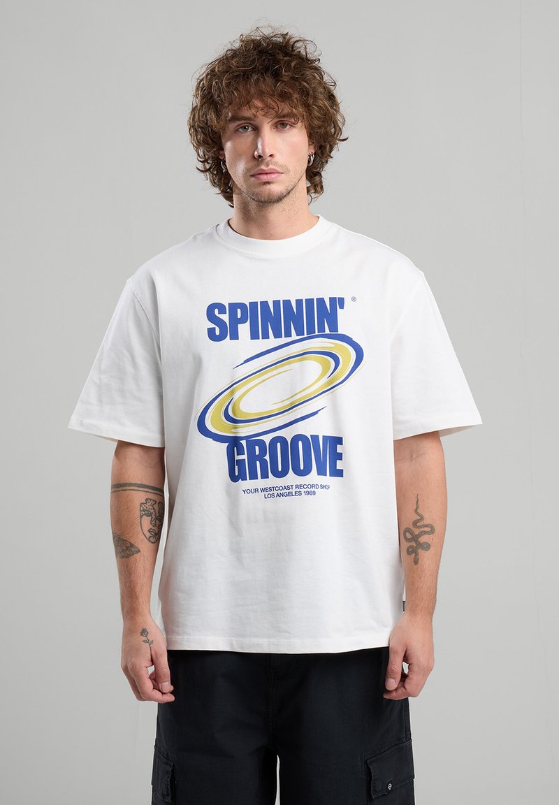 T-shirt en coton blanc avec un grand design graphique bleu et jaune comportant le texte "SPINNIN' GROOVE" et un motif circulaire. Manches courtes, coupe décontractée.