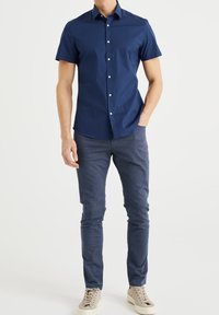 Muž v námořnické modré košili na krátký rukáv, těsných modrých kalhotách a světle béžových teniskách, s jednou rukou v kapse na bílém pozadí.