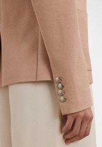 Blazer texturé rose clair avec quatre boutons métalliques sur les poignets. Le tissu est tissé, présentant un motif subtil. Conception ajustée.
