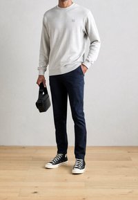 Szary sweter, granatowe spodnie i czarne sneakersy; trzyma małą czarną torbę. Prosty design z długimi rękawami i elastycznymi mankietami.