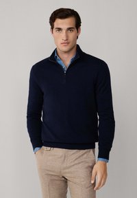 Mann trägt einen navyblauen Viertelzip-Pullover über einem hellblauen Hemd, kombiniert mit beigen Hosen, vor einem schlichten grauen Hintergrund.