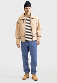Veste en polaire beige avec des accentuations marron, chemise rayée en dessous, jeans bleus et bottes beiges. Stylé avec un bonnet noir.