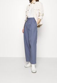Persona con pantaloni blu a vita alta, top bianco a coste, giacca beige e sneakers bianche in piedi davanti a uno sfondo bianco uniforme.