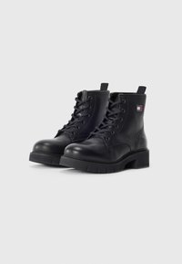 Bottines noires en cuir avec un design à lacets, semelles épaisses, et petites languettes. Présente un écusson logo sur le côté.