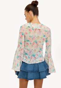 Blouse fleurie en tissu transparent présentant des couleurs pastel, des manches longues évasées, un col et un ourlet volantés, associée à une jupe en jean à volants.