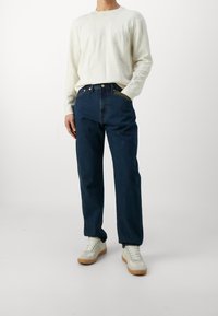 Personne portant un pull à manches longues blanc, un jean bleu foncé à jambes droites et des baskets blanches à semelles marron, debout avec une main dans la poche.