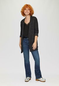 Lange, zwarte, gebreide cardigan met opgerolde mouwen over een zwarte top. Gemaakt met blauwe flared jeans en zilveren klompen, met een houtachtige zool.