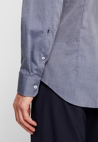 Niebieska koszula z długim rękawem z teksturowanym wzorem, białymi zapięciami na mankietach oraz zaokrąglonym dołem. Zaprojektowana z myślą o dopasowanym kroju.