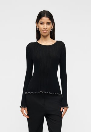 Kvinde med langt mørkt hår iført sort ribstrikket langærmet top med flæsekanter ved ærmer og kant, kombineret med sorte bukser, står foran en ensfarvet baggrund.