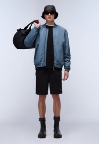 Lichtblauwe bomberjack met elastische manchetten, zwart t-shirt, zwarte shorts, zwarte sokken en zwarte laarzen. Model houdt een zwarte sporttas vast.