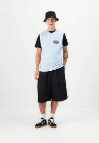 Ung man i svart bucket hat, ljusblå ärmlös skjorta över svart t-shirt, svarta vida shorts, vita strumpor och svarta sneakers stående mot vit vägg.